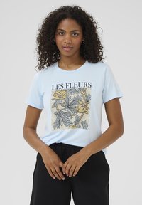 Lichtblauw T-shirt met korte mouwen en ronde hals, voorzien van een bloemenafdruk met "LES FLEURS" in vetgedrukte zwarte tekst.