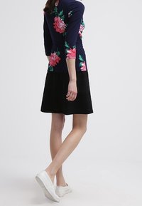 Marinblus med blommönster och trekvartslånga ärmar, kombinerad med en svart kjol. Vita sneakers fulländar outfiten, vilket ger en avslappnad och figurnära design.