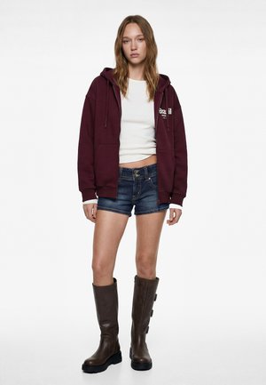 Joven mujer vistiendo una sudadera con capucha burdeos con cremallera, top blanco corto, pantalones cortos de mezclilla y botas altas de cuero marrón, posando frente a un fondo blanco.