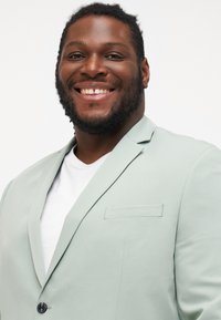 Χαμογελαστός άνδρας με κοντά σγουρά μαλλιά και γένια φορεί ανοιχτό γκρι σακάκι πάνω από λευκό πουκάμισο, ποζάροντας μπροστά σε απλό λευκό φόντο.