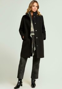 Abrigo de lana negra con cuello alto, botones plateados, combinado con pantalones oscuros estampados y una bufanda en capas verde y gris. Textura suave, ajuste entallado.