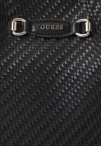 Texture en cuir tissé noir avec une sangle en cuir noir ornée de ferrures argentées et le mot « GUESS » embossé en lettres argentées.