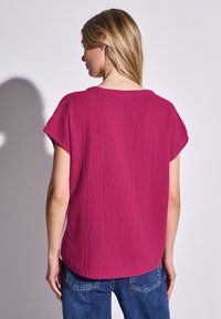 Top a maniche corte, leggero e morbido, di colore fucsia con una finitura testurizzata. Collo rotondo e orlo leggermente curvato. Indossato con jeans blu.