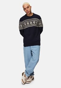 Navy blue knitted sweater with white and cream geometric pattern across the chest, paired with light blue jeans and beige sneakers.
