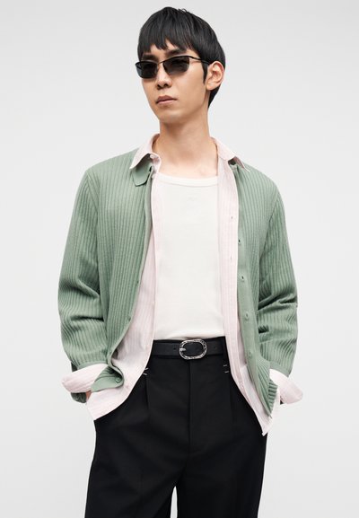 Ανοιχτό πράσινο ριμπ κασκόλ πάνω από ένα ροζ ριγέ πουκάμισο, φορεμένο με λευκό τοπ χωρίς μανίκια και μαύρο παντελόνι, με μαύρη ζώνη με ασημένια αγκράφα.