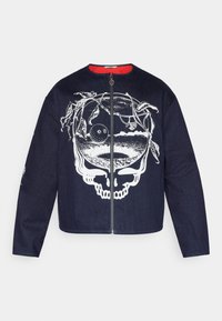 Veste en denim foncé avec un grand motif de crâne blanc orné de vignes. Fermeture éclair frontale, doublure intérieure rouge, manches longues.