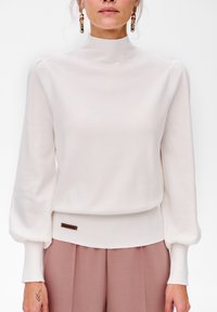 Biała sweter z wysokim kołnierzem, długimi puff sleeves, prążkowanymi mankietami i dopasowanym dołem. Posiada małą brązową metkę w pobliżu dolnej krawędzi.