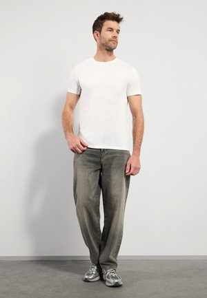Weißes T-Shirt mit kurzen Ärmeln, kombiniert mit lockeren grauen Jeans. Das Model trägt hellgraue Sneakers und steht auf einem strukturierten grauen Boden.