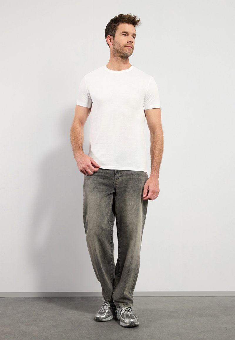 T-shirt blanc à manches courtes associé à un jean gris ample. Le mannequin porte des baskets gris clair et se tient sur un sol texturé gris.