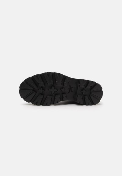 Semelle de chaussure en caoutchouc noir avec des rainures profondes et des motifs en forme de croix pour l’adhérence, présentée sur un fond blanc uni.