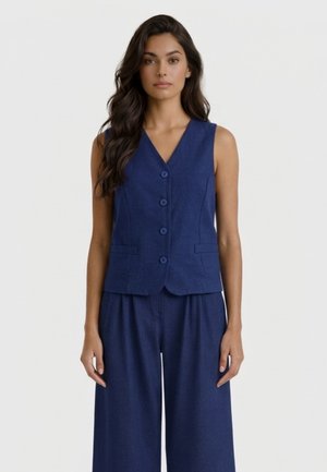 Donna con capelli lunghi e scuri indossa un gilet blu navy senza maniche con bottoni e pantaloni larghi abbinati, in piedi davanti a uno sfondo chiaro e uniforme.