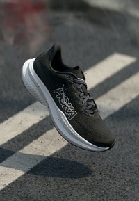 Chaussure de sport noire avec une tige texturée, un bout renforcé et un grand logo "Hoka" blanc. Elle dispose de lacets gris et d'une épaisse semelle blanche.