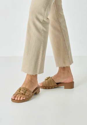 Sandales à enfiler tressées marron avec un petit talon bloc, ornées d'une boucle dorée et d'accents cloutés le long de la semelle.