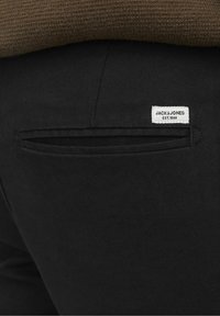 Gros plan sur la poche arrière d'un pantalon noir avec une étiquette blanche JACK & JONES et une personne portant un pull marron texturé.