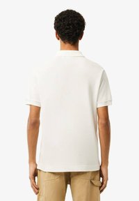 Polo bianco con una consistenza morbida, maniche corte e colletto a maglia. La vista dal retro mostra una finitura liscia senza branding o motivi visibili.