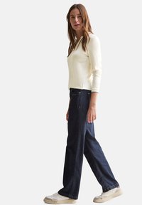 Top crema a maniche lunghe con scollatura rotonda, abbinato a jeans larghi blu scuro e sneakers bianche con accenti beige.