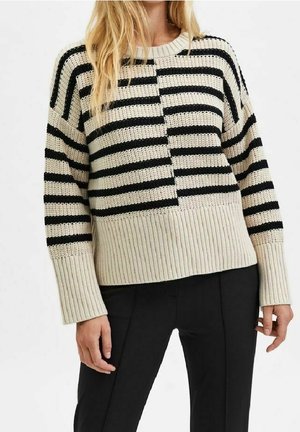Femme portant un pull en maille beige avec des rayures horizontales noires et un pantalon noir, vue du cou aux hanches.