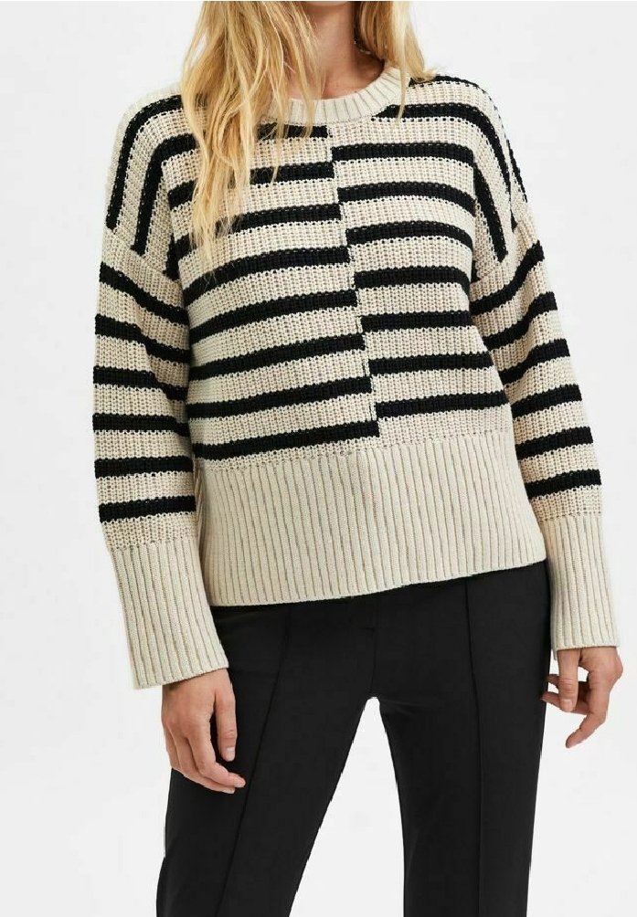 Femme portant un pull en maille beige avec des rayures horizontales noires et un pantalon noir, vue du cou aux hanches.