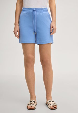 Frau trägt hellblaue Shorts mit Kordelzug und beige Slides mit schwarzem Text, steht vor einem schlichten hellen Hintergrund.