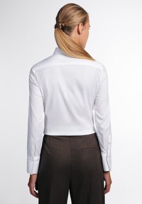 Camicia bianca a maniche lunghe con colletto classico e polsini abbottonati, abbinata a pantaloni marroni su misura. Tessuto liscio, linee pulite, aspetto professionale.