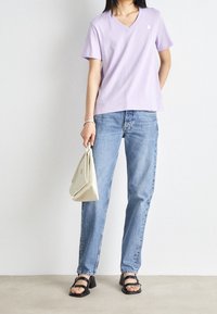 Lila V-ringad t-shirt, kortärmad, parat med ljusblå raka jeans. Håller en krämfärgad handväska, bär svarta sandaletter med remmar.