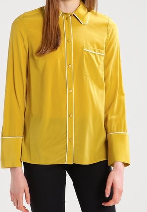 Donna che indossa una camicia giallo senape con bottoni e dettagli di piping bianchi e pantaloni neri su uno sfondo uniforme.