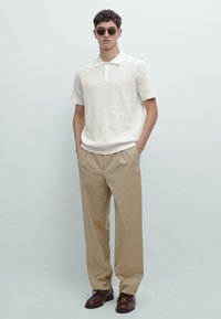 Polo a maglia color beige chiaro con colletto, abbinata a pantaloni kaki ampi e scarpe di pelle marrone. Occhiali da sole rotondi completano il look.