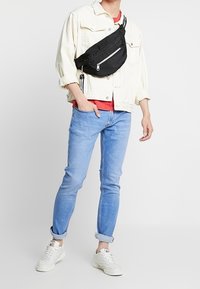 Bolso cruzado negro con bolsillos con cremallera, usado sobre una chaqueta de denim blanca, camisa roja, jeans azules y zapatillas blancas. Conjunto casual.