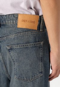Denim jeans in vervaagde blauw met een leren label in bruin op de tailleband, voorzien van twee achterzakken en zichtbare stikdetails.