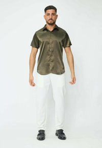 Chemise en satin vert olive à manches courtes avec un col pointu, fermeture à boutons et ourlet arrondi, associée à un pantalon blanc et des chaussures noires.