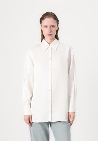 Camicia bianca a maniche lunghe con colletto classico. Tessuto morbido con dettagli in rete sui lati. Indossata con pantaloni di jeans azzurri chiari.