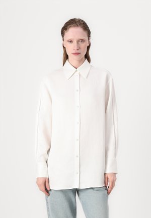 Camicia bianca a maniche lunghe con colletto classico. Tessuto morbido con dettagli in rete sui lati. Indossata con pantaloni di jeans azzurri chiari.