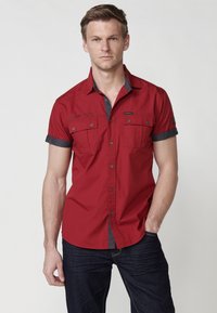 Camisa de manga corta roja con detalles en gris, frontal abotonado, dos bolsillos en el pecho y cuello en contraste. Textura suave y diseño ajustado.