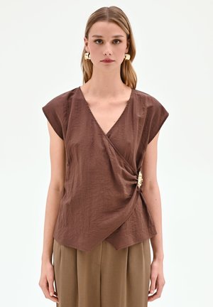 Femme portant un top croisé marron à manches courtes avec une boucle dorée sur le côté et un pantalon ample beige, debout devant un fond uni.