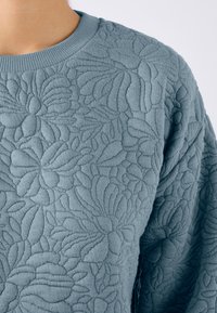 Suéter azul acolchado con textura floral, que presenta un cuello redondo acanalado y una tela suave. El diseño incluye patrones prominentes de hojas y flores.