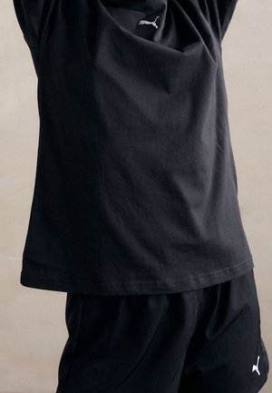 Osoba ubrana w czarną koszulkę Puma i spodenki, unosząca ramiona na neutralnym tle.