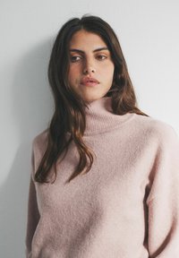 Pehme roosa kõrge kaelusega kampsun, valmistatud tekstuurse koo kangast, lõdva lõikega ja soonilise kraega, eksponeeritud lihtsa tausta ees.