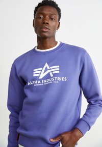 Vijoličast pulover iz mešanice bombaža, ki ima spredaj bel logo "Alpha Industries" in grafični dizajn, okroglo izreznico in rebraste manšete.