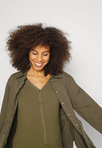 Camisa olive green de botão, com uma parte de cima ribada em olive green por baixo, apresentando um decote em V. Textura suave, corte descontraído e mangas largas.