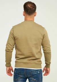 Uomo con capelli castani corti che indossa un felpa beige e jeans blu strappati, in piedi con le spalle rivolte verso l'osservatore su uno sfondo neutro.