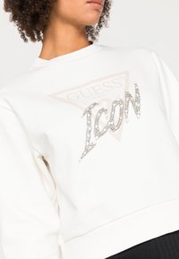 Valkoinen, pyöreäkauluksinen collegepaita, jossa on suuri vaaleanpunertava logo ja "ICON" hopeisissa paljeteissa. Pehmeää kangasta, jossa ribbineulotut hihansuut ja helma.