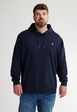 Sudadera de color azul marino con un bolsillo frontal, cordones y un pequeño logo. Tejido suave y corte holgado, combinada con jeans de mezclilla azul.