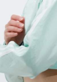 Svetlo tyrkysová vetrovka s bielymi pruhmi pozdĺž rukávov, ktorá sa vyznačuje hladkou textúrou a uvoľneným strihom. Ruka je čiastočne viditeľná, ako pevne drží látku.