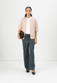 Light pink, short-sleeved cardigan with button closure, paired with dark green wide-leg pants, and holding a small black bag. Black shoes.