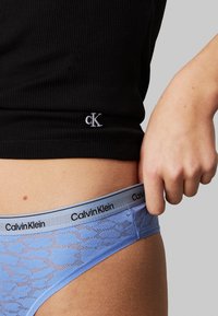 Šviesiai mėlynas nėriniuotasis tangas su juodu Calvin Klein juosmeniu. Detaliame plane matoma tekstūruota medžiaga ir ranka, suimanti juosmenį.