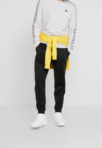 Šedá dlouhá tričko s černými bočními pruhy, černé tepláky a žlutá mikina uvázaná v pase. Bílí tenisky dotvářejí vzhled.