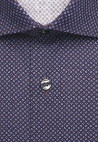 Gros plan sur un col de chemise marron avec de petits motifs losanges bleus et un seul bouton gris fermé près du cou.