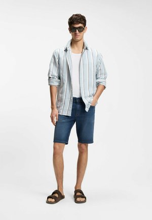 Junger Mann mit schwarzen Sonnenbrillen, hellblau-weiß gestreiftem Hemd über einem weißen T-Shirt, Jeansshorts und braunen Sandalen, steht mit einer Hand in der Tasche.