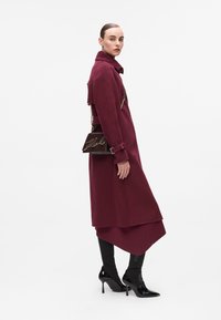 Burgunderfarbener Wollmantel mit Kragen, taillierter Taille und langen Ärmeln; kombiniert mit schwarzen Stiefeletten und einer braunen Handtasche mit Kettenriemen.