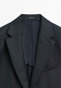 Blazer grigio scuro con rever a punta, tessuto strutturato, foderato all'interno e una tasca sul petto. L'etichetta reca la dicitura "Made in Portugal."